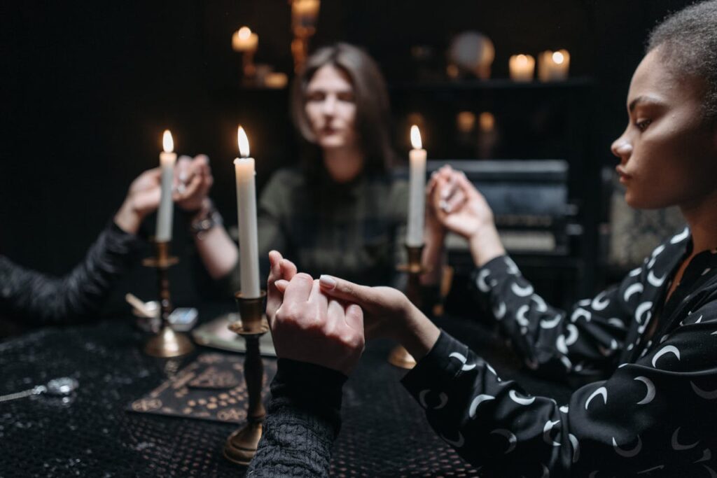 A mystical seance with three women holding hands around candlesticks in a dimly lit room.