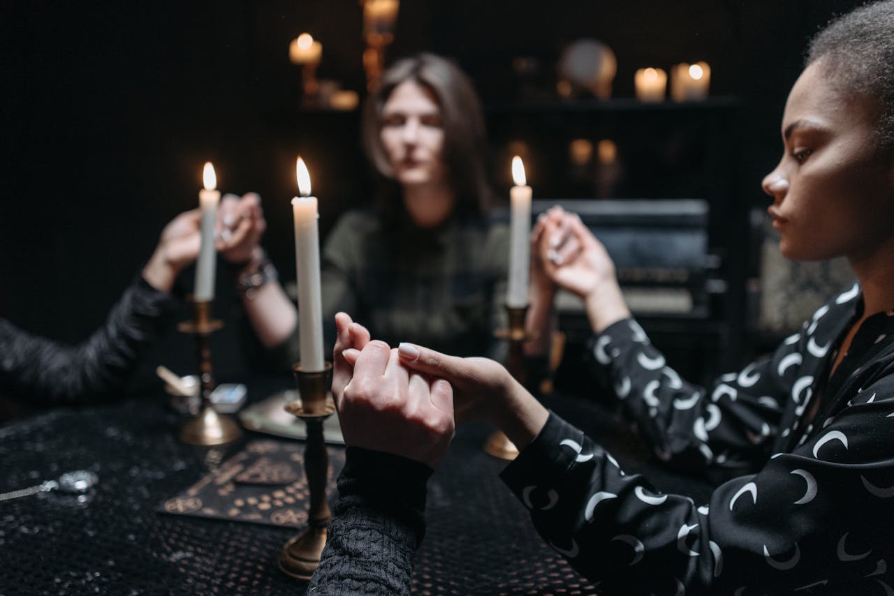 A mystical seance with three women holding hands around candlesticks in a dimly lit room.